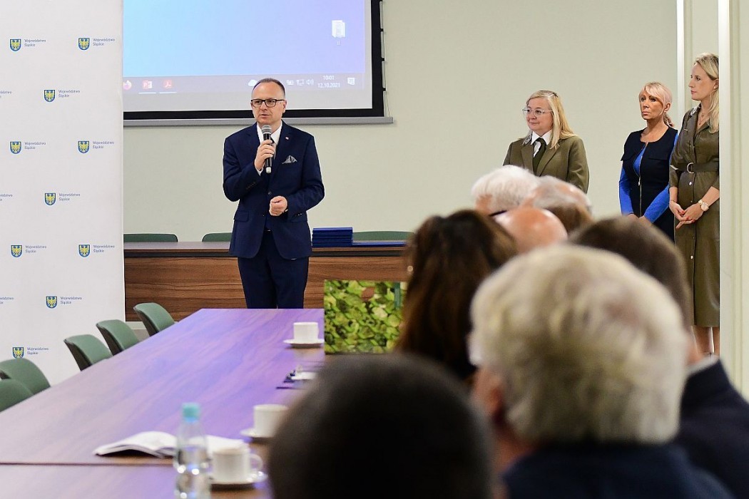 Mężczyzna z mikrofonem przemawiający do osób siedzących przy stole. Obok prowadzącego stoją trzy kobiety. W tle baner Województwa Śląskiego.