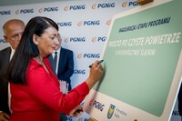 Kobieta w czerwonej bluzce podpisująca się na plakacie z napisem inauguracja I etapu programu prosto po czyste powietrze w Województwie Śląskim. Obok niej stoi dwóch mężczyzn na tle baneru z logo PGNiG.