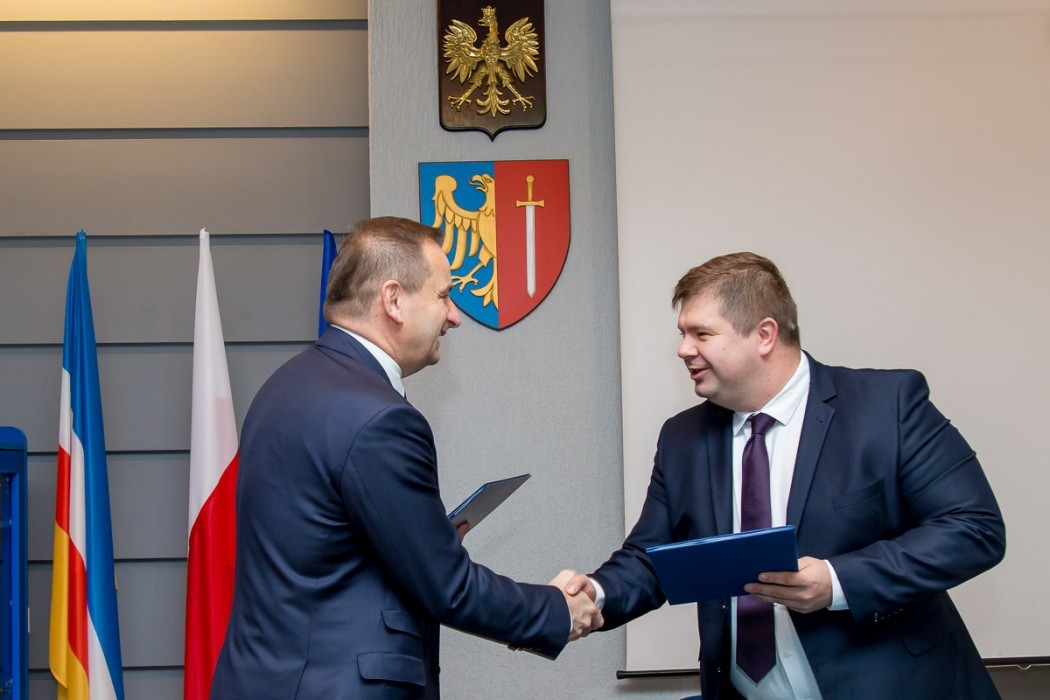 Mężczyźni ściskający sobie dłonie i trzymający teczki. W tle flagi oraz wiszące godło i herb.