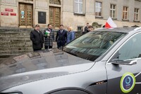 Mężczyźni i kobieta stojący przed gmachem Urzędu Marszałkowskiego. Przed nimi stoi samochód. w tle widać flagę Polski.