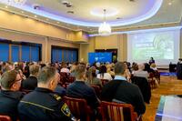 Sala konferencyjna z uczestnikami siedzącymi na krzesłach, skierowanymi w stronę dużego ekranu z prezentacją zatytułowaną ‘Konferencja’; w tle widoczne niebieskie tablice informacyjne, żyrandole i oświetlenie sufitowe.