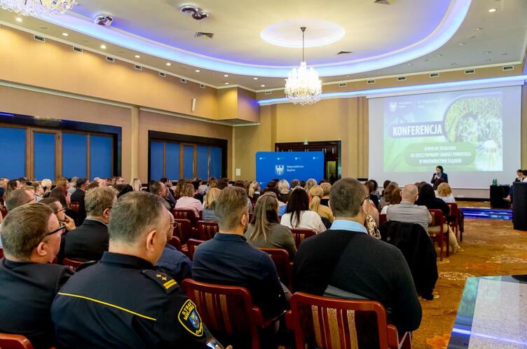 Sala konferencyjna z uczestnikami siedzącymi na krzesłach, skierowanymi w stronę dużego ekranu z prezentacją zatytułowaną ‘Konferencja’; w tle widoczne niebieskie tablice informacyjne, żyrandole i oświetlenie sufitowe.