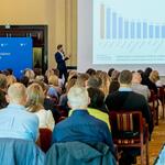 Sala konferencyjna z uczestnikami, Piotr Łyczko – Zastępca Dyrektora Departamentu Ochrony Powietrza i Negocjacji Klimatycznych w Ministerstwie Klimatu i Środowiska prezentuje zmiany wynikające z dyrektywy AAQD i ich wpływ na programy ochrony powietrza; w tle niebieska ścianka z herbem województwa