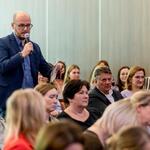 Uczestnik konferencji z mikrofonem zadaje pytanie, w tle widoczni pozostali uczestnicy siedzący w sali.” 