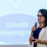 Karolina Pancewicz-Kosno z referatu ds. planowania w zakresie środowiska w Departamencie Ochrony Środowiska, Ekologii i Opłat Środowiskowych Urzędu Marszałkowskiego Województwa Śląskiego prowadzi prelekcję trzymając mikrofon i pilot do slajdów, w tle ekran z napisem ‘Uchwała antysmogowa’