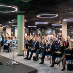 Prelegent w garniturze przemawia przy szklanej mównicy, za nim ekran z jego wizerunkiem i zielonym tłem roślinnym