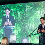 Wojewoda Śląski przemawia przy mównicy, za nim duży ekran z jego wizerunkiem i zieloną dekoracją roślinną