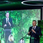 Członek Zarządu Województwa Śląskiego przemawia przy mównicy, za nim duży ekran z jego wizerunkiem i zieloną dekoracją roślinną