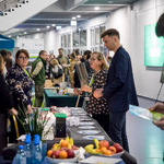 Stoisko informacyjne z materiałami promocyjnymi i owocami na stole, uczestnicy rozmawiają w przestrzeni targowej