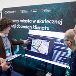 Prezentacja na stoisku „Coolcity” z dużym ekranem pokazującym mapę miasta, uczestnicy oglądają i dyskutują