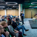 Prelegent przemawia na scenie do publiczności, obok stoi osoba z notatkami, w tle ekran z prezentacją