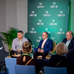 Panel dyskusyjny w sali konferencyjnej. Pięć osób siedzi na scenie na krzesłach, przed nimi stoliki z wodą. W tle zielona ścianka z logo „CLIMATE-CON” powtarzającym się w regularnym wzorze. Po lewej stronie osoba moderująca trzyma kartki. Na pierwszym planie widać kilka krzeseł dla publiczności