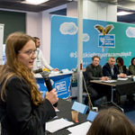 Osoba stojąca przy stole trzyma mikrofon i przemawia podczas debaty. Na stole leżą kartki, kubki i tabliczki z nazwiskami. W tle widoczny baner z napisem „Zespół Szkół Licealnych i Technicznych w Rydultowach” oraz duża niebieska plansza z hasłem „Śląskie Przywracamy Błękit”. Za stołem siedzi kilka osób w formalnych strojach