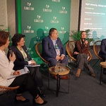 Panel dyskusyjny z sześcioma osobami siedzącymi na scenie na krzesłach z wiklinowymi oparciami. Na stolikach stoją szklanki z wodą. W tle zielona ścianka z logo „CLIMATE-CON” oraz ekran z agendą sesji. Na ekranie widoczne są napisy dotyczące strategii i tematów rozmowy