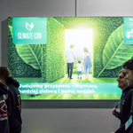 Grupa osób stoi przed dużym ekranem LED z grafiką przedstawiającą rodzinę wchodzącą do zielonego korytarza z liści. Na ekranie widoczny napis „CLIMATE-CON” oraz hasło „Budujemy przyszłość – czystszą, bardziej zieloną i pełną nadziei”
