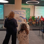 Dwie osoby stoją przy sztaludze i rysują kolorowe elementy na dużej planszy. W tle scena z panelem dyskusyjnym – pięć osób siedzi na krzesłach, za nimi zielona ścianka z logo „CLIMATE-CON” i dwa ekrany z prezentacją. Na ekranach widoczne są napisy dotyczące strategii dla województwa