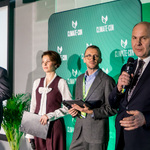 Cztery osoby stoją na scenie, jedna przemawia do mikrofonu, w tle plansza „Climate-Con”