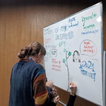 Osoba rysuje na dużej białej tablicy suchościeralnej kolorowe ilustracje i napisy. Na tablicy widoczne są słowa „ŚLĄSK”, „Rzeszów”, „CO2 out”, „Fajne projekty warsztatów” oraz rysunek drzewa. Obok stoi druga osoba, która pomaga przy tworzeniu grafiki